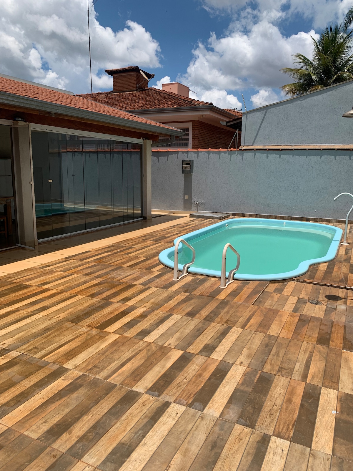 A private outdoor area features a rectangular pool with steps for easy access, surrounded by a wooden deck. Glass sliding doors lead to an adjacent indoor space, while a concrete wall provides a sense of privacy. Lush greenery can be seen in the background.