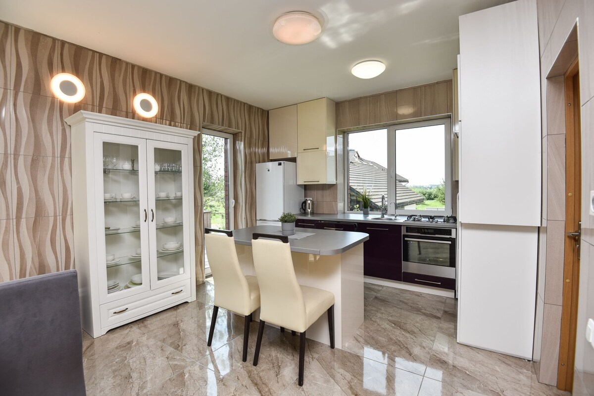 A modern kitchen is presented with sleek cabinetry and a culinary island. Two light-colored chairs are positioned at the island, while large windows invite natural light. A glass-fronted cabinet displays dining essentials, and appliances are integrated for convenience.