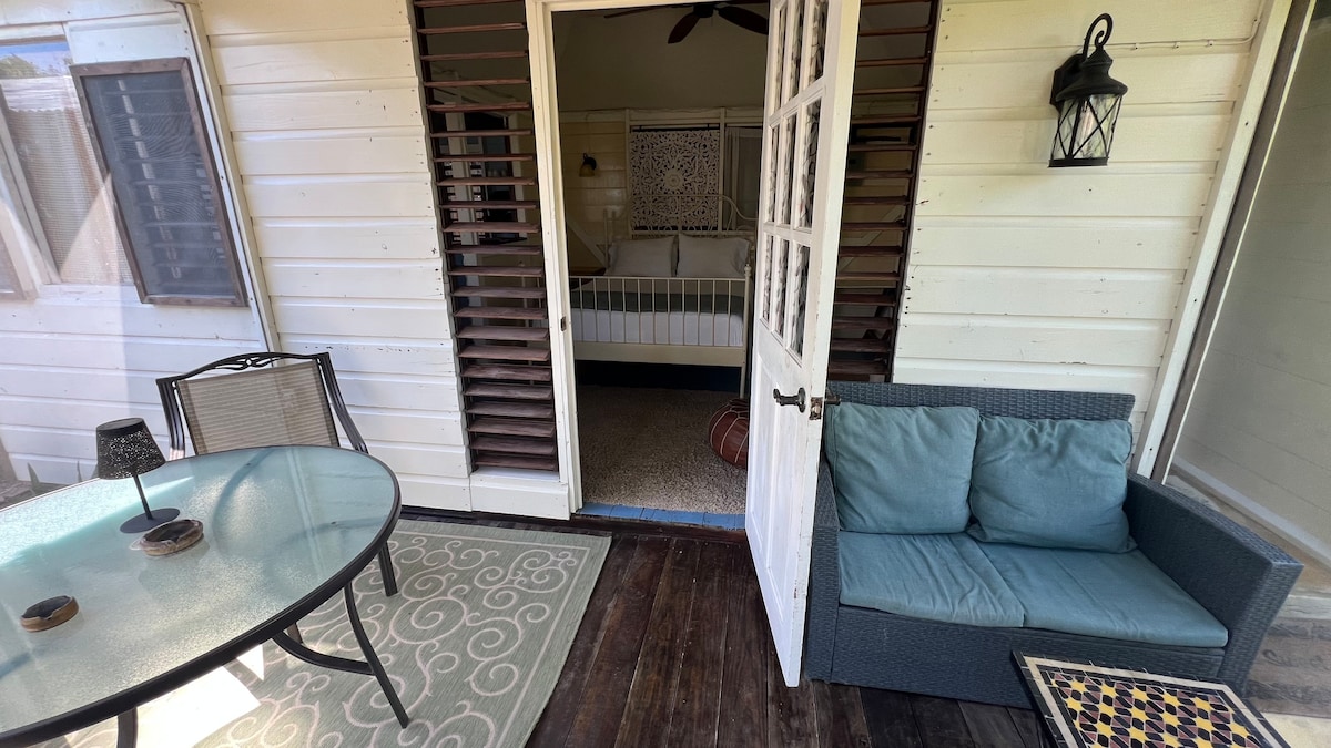 A cozy entrance leads to a shaded patio featuring a round glass table and black chairs. A comfortable blue seating area invites relaxation. Beyond, an open door reveals a bedroom with a light-colored bed, adding warmth to the space, while natural light filters through the windows.