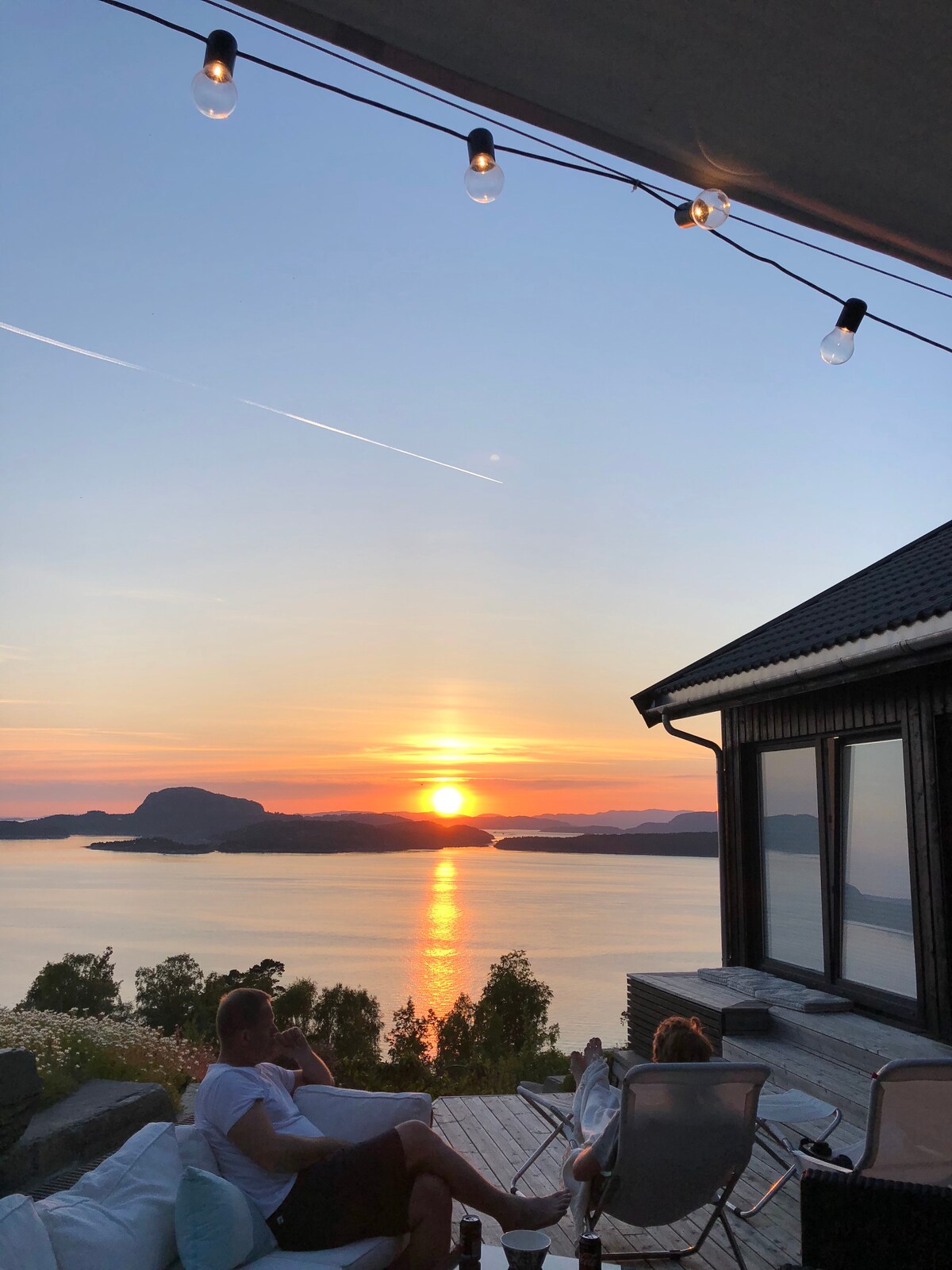 A stunning sunset view over the calm waters, with warm tones reflecting on the surface. A deck furnished with comfortable seating is visible, and a cozy home is positioned to the side. String lights provide a soft glow in the evening ambiance.