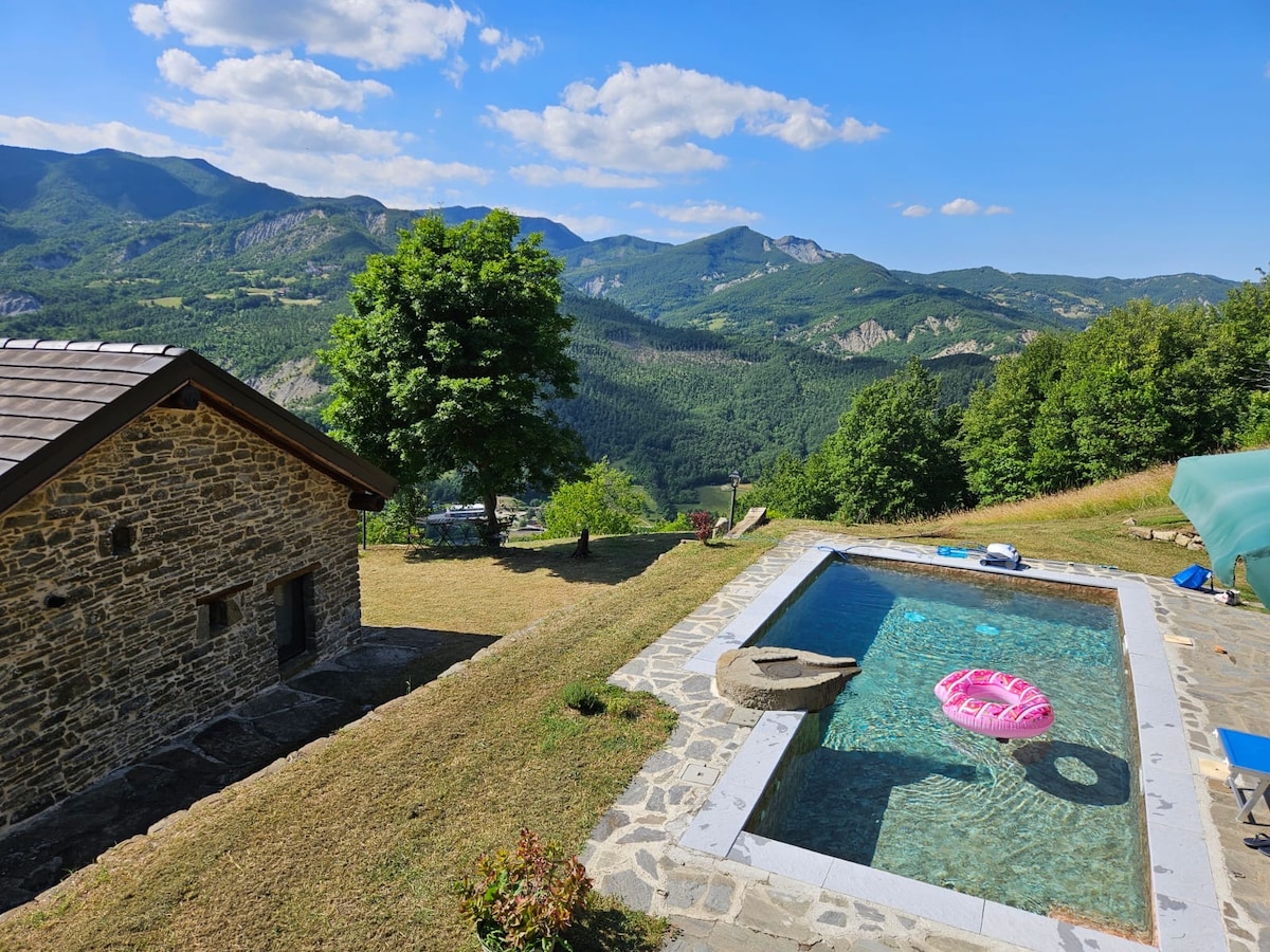 A rustic stone cottage is framed by lush greenery, with a serene swimming pool positioned nearby. The pool area features a floating lounge and offers expansive views of rolling hills and mountains under a clear blue sky, enhancing the natural surroundings.