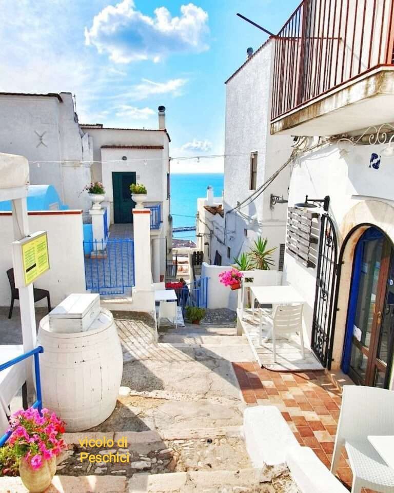 A narrow street is depicted, lined with whitewashed buildings. Potted plants and colorful flowers enhance the entrance of a café. Tables and chairs are arranged on a cobbled path that leads toward a glimpse of the sea in the distance, reflecting the sky.