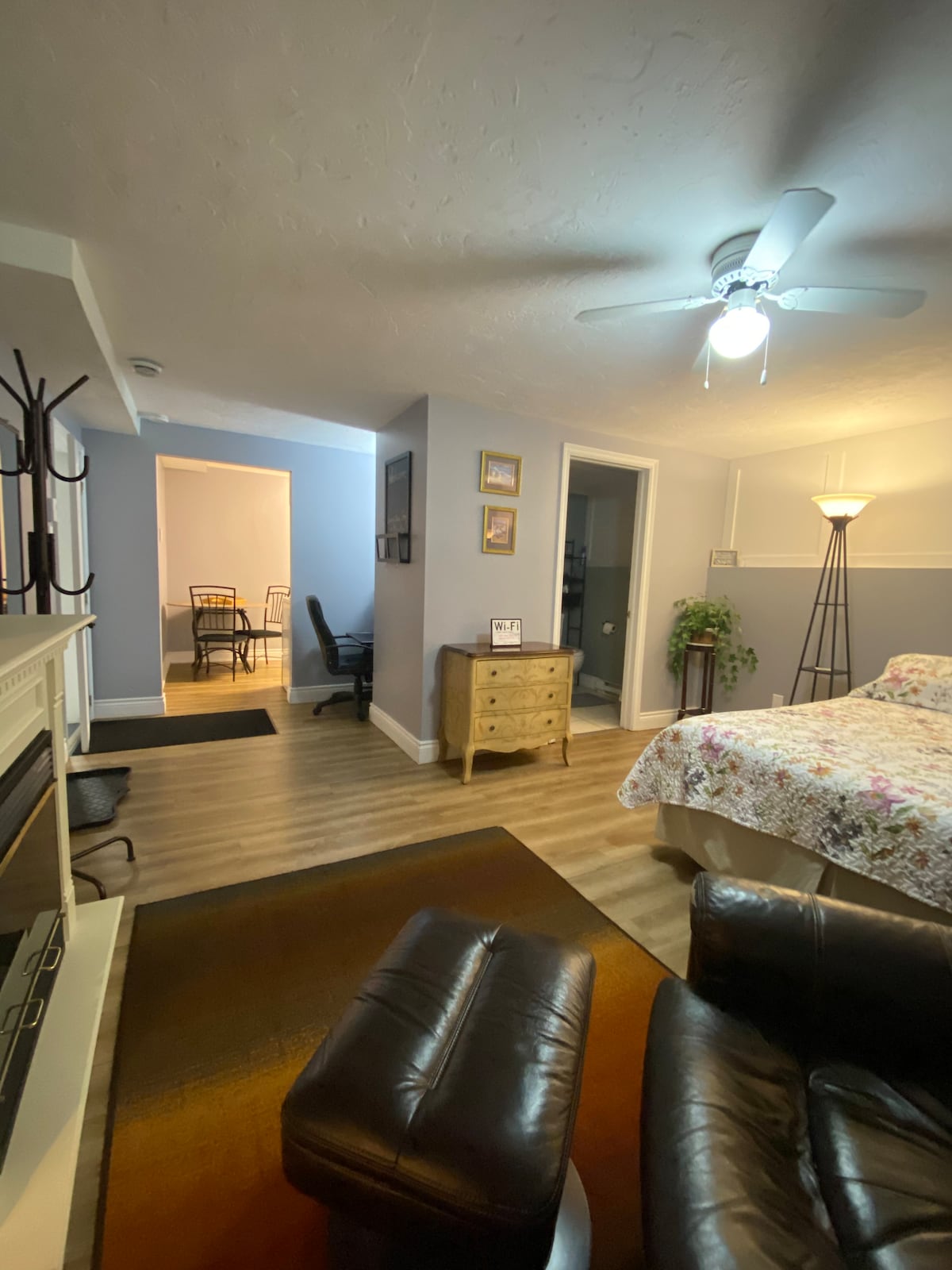 The room showcases a spacious layout, featuring a decorative fireplace and a cozy seating area with leather chairs. A ceiling fan provides airflow above a floral-patterned bed. Natural light brightens the space, complemented by a standing lamp and visible dining area in the background.