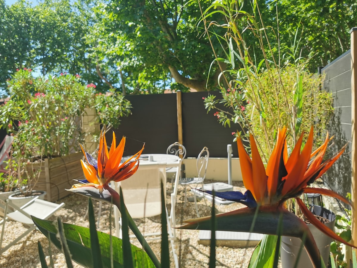 A serene outdoor space is featured, showcasing vibrant orange and blue bird of paradise flowers in the foreground. In the background, a small dining table with chairs is surrounded by lush greenery, creating a peaceful atmosphere in the private courtyard.