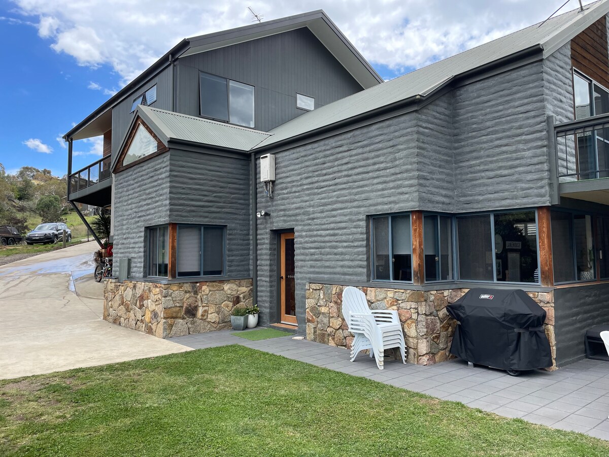 An exterior view of the Lakefront@Tyrolean house showcases its modern design with a combination of stone and textured siding. A spacious outdoor area features a patio with seating, a charcoal grill, and manicured green lawns, providing an inviting setting for gatherings.