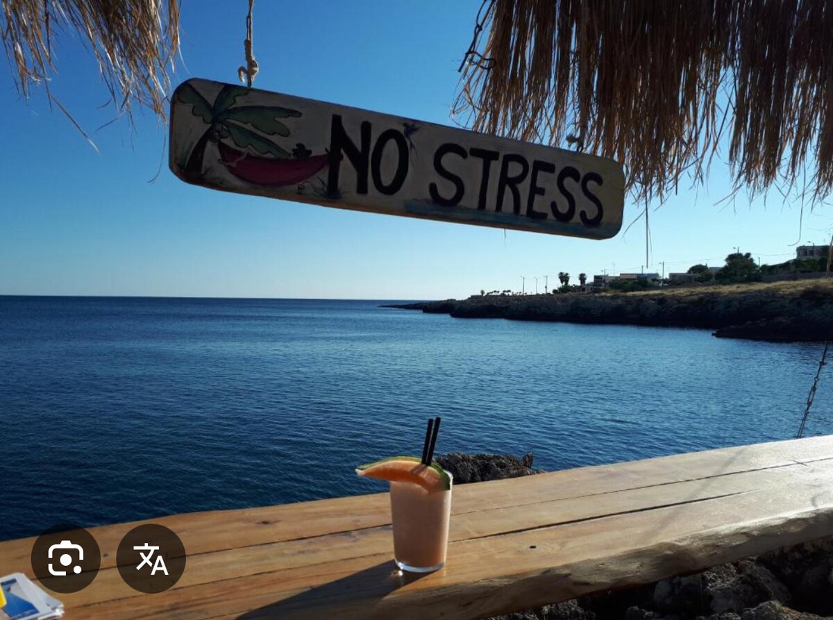 A wooden sign featuring the phrase 'NO STRESS' hangs above a rustic table. A refreshing cocktail, garnished with a slice of fruit, rests on the table. In the background, calm blue waters meet a clear sky, with a hint of coastal scenery visible.