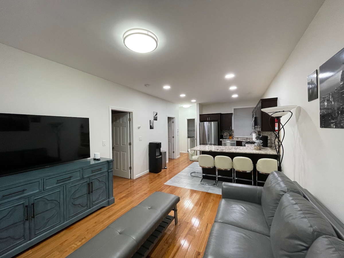 The open-plan living area features a comfortable gray sofa facing a large flat-screen TV. The adjoining kitchen showcases modern cabinetry, a spacious countertop, and seating for four at the bar. Warm wood flooring runs throughout, enhancing the cozy ambiance of the space.