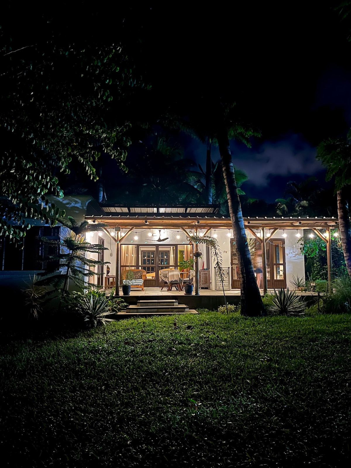 The retreat is illuminated at night, highlighting its welcoming exterior. The covered patio features wooden furniture and is framed by tropical plants. Soft lighting enhances the peaceful ambiance, while the surrounding garden is visible under the starlit sky.