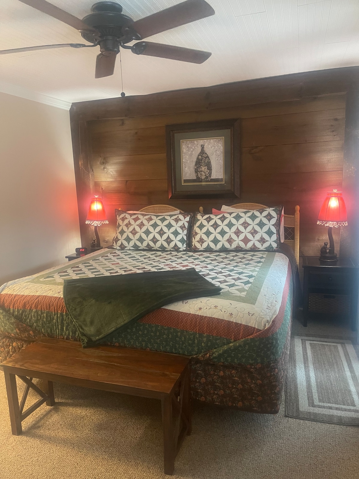 A cozy bedroom features a spacious bed with a patchwork quilt and decorative pillows, complemented by a wooden accent wall. Two bedside lamps provide soft lighting, and a small bench is positioned at the foot of the bed, adding functional space.