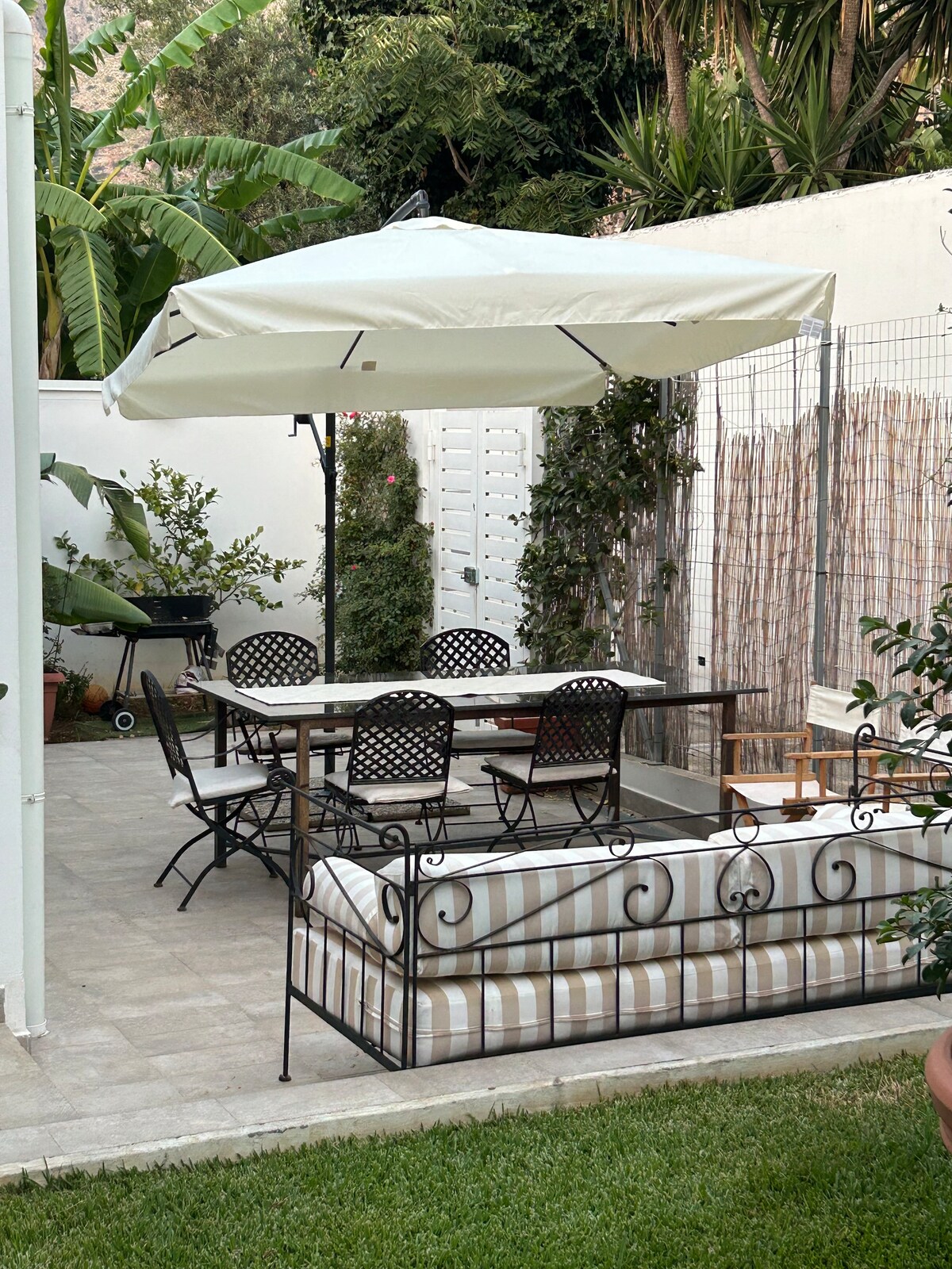 An outdoor terrace features a dining table with six chairs under a white umbrella. A comfortable sofa with striped upholstery is positioned nearby, alongside greenery and palm trees creating a serene garden setting.