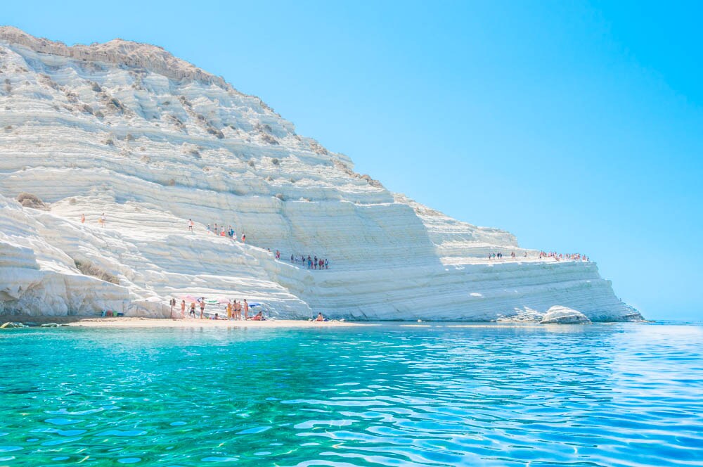 A rocky coastline is featured, characterized by striking white cliffs contrasting with the deep blue sea. Sunbathers can be seen on the beach below, while gentle waves lap at the shore, creating a serene setting filled with natural beauty.