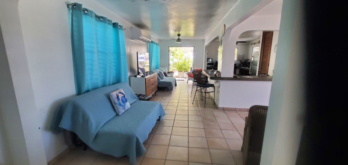 A spacious living area features light blue curtains that filter natural light, with cozy seating options including two sofas. A dining area with bar seating is visible, alongside a well-equipped kitchen in the background. Tiled flooring extends throughout the room.