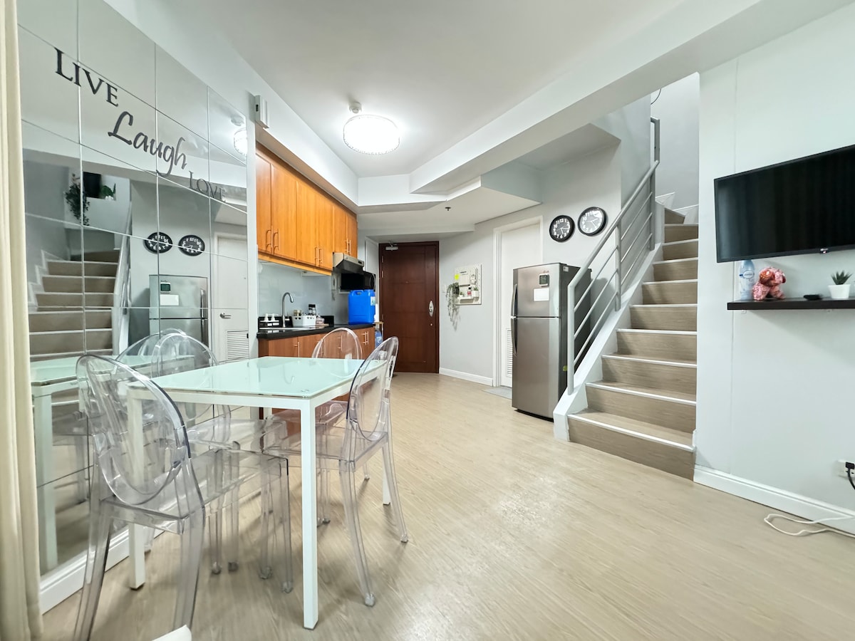 A modern dining area is featured with a glass table and transparent chairs. The kitchen is fully equipped with wooden cabinetry, a refrigerator, and appliances. A staircase leads to the upper level, while large mirrors enhance the sense of space and light in the unit.