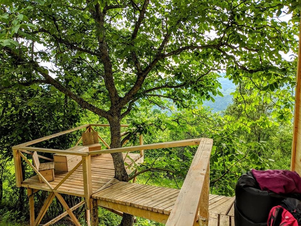 A wooden terrace is nestled among lush green trees, overlooking a valley. The structure features natural wood railings and a sturdy floor, creating a peaceful outdoor space. Framing the scenic view, foliage adds a sense of tranquility to the surroundings.