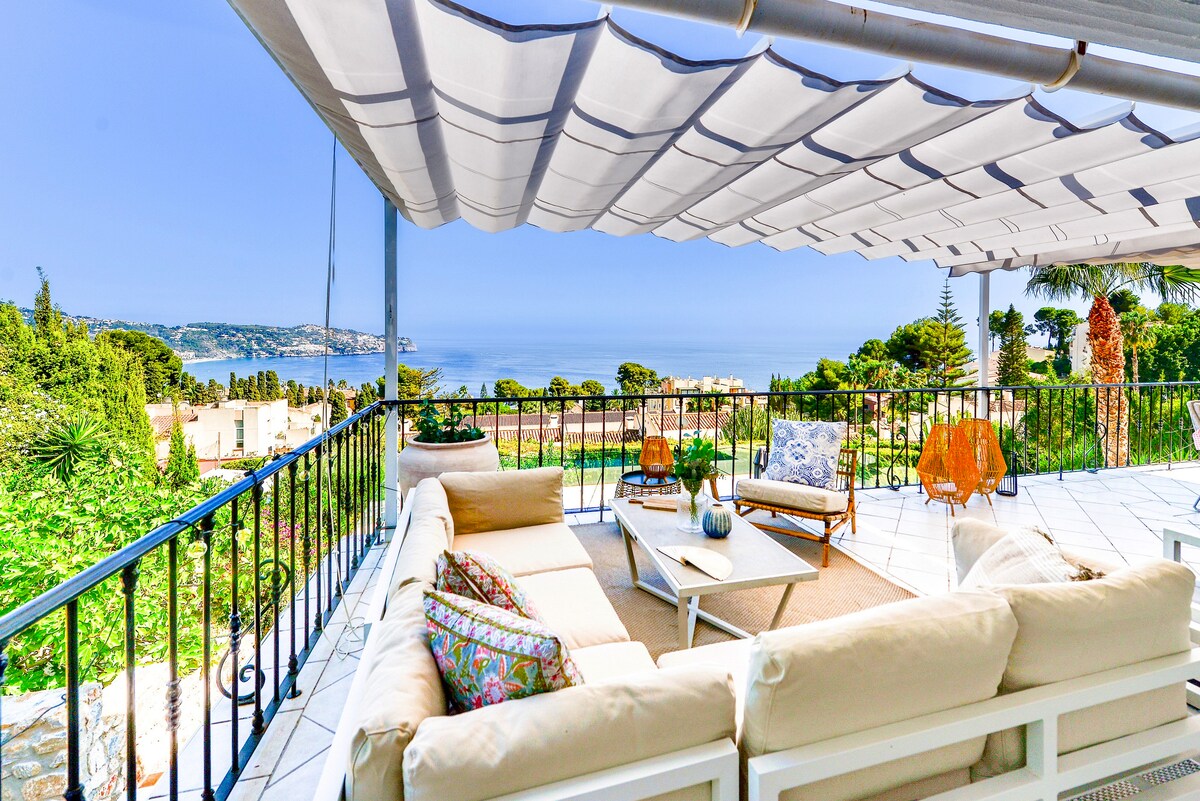 A spacious terrace features a modern lounge sofa adorned with colorful cushions, inviting relaxation. A coffee table sits centrally, surrounded by lush greenery and a panoramic view of the La Herradura bay. Partially shaded by a canopy, the area is designed for comfortable outdoor enjoyment.