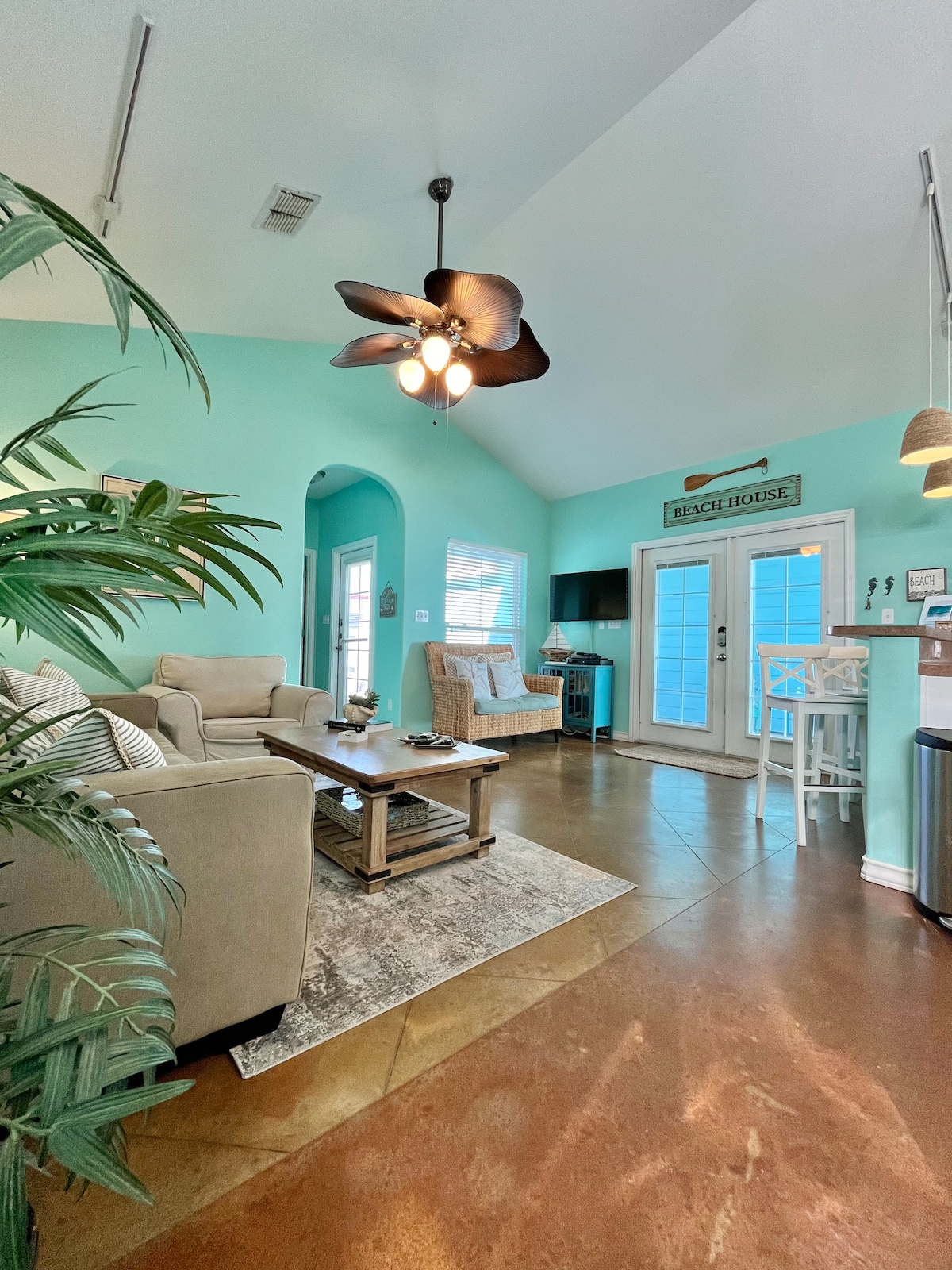 A bright living area features walls painted in a serene turquoise hue. Natural light fills the space through large windows. Comfortable seating is arranged around a wooden coffee table, while a ceiling fan and decorative elements enhance the coastal vibe. Access to an outdoor area is provided through glass doors.