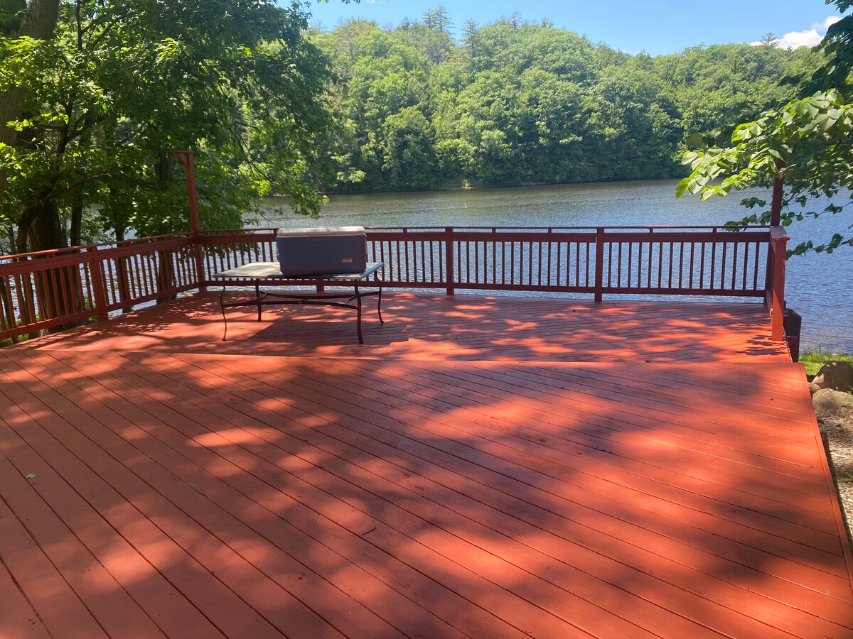A spacious red deck overlooks a calm river, framed by lush greenery and trees. The sunlight casts playful shadows on the deck surface, creating a serene setting for relaxation or outdoor activities by the water.