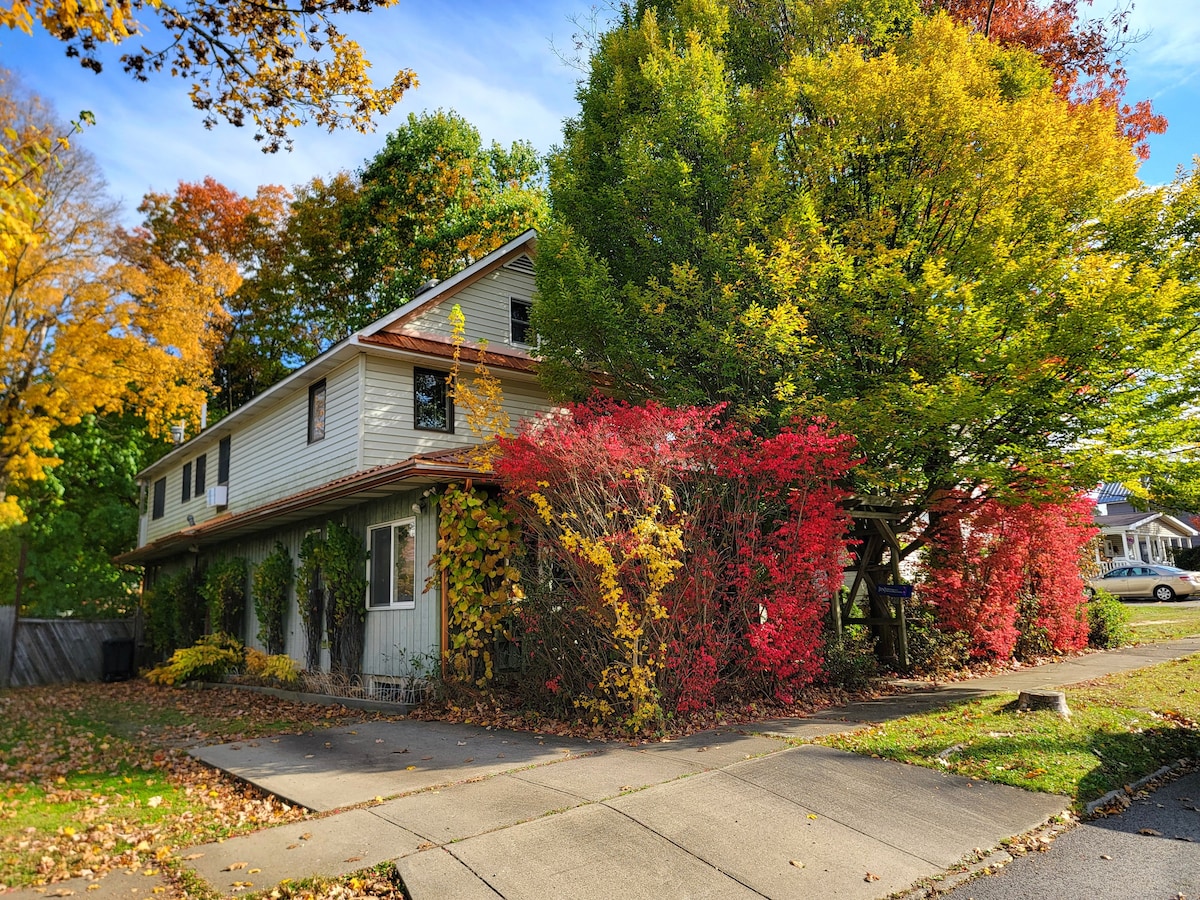 The exterior of a two-story home is surrounded by vibrant fall foliage, featuring rich reds, greens, and yellows. Lush trees and colorful shrubs frame the entrance, while a paved pathway leads to the house. The sky above is clear, enhancing the warm seasonal colors.