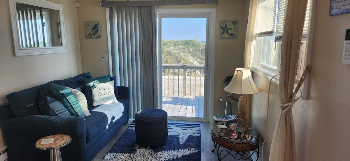 A welcoming sitting area features a navy blue sofa adorned with decorative pillows, facing a patio door that leads to a partial ocean view. A circular ottoman is placed on a star-patterned rug, while a lamp and a small side table provide additional comfort.