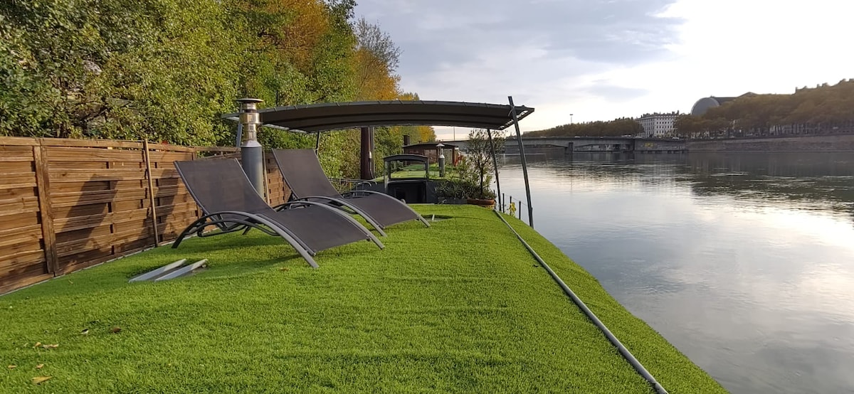 A serene outdoor area of the houseboat features two lounge chairs positioned on a grassy surface. A shaded pergola provides cover, while the calm river reflects trees along the bank. The peaceful setting offers a direct view of the waterway.