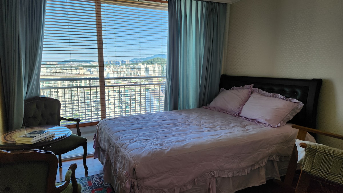 A well-lit bedroom features a spacious bed with light-colored bedding, positioned near a large window that offers a view of the surrounding cityscape. A small table with books and a comfortable chair, along with fresh curtains, enhance the inviting atmosphere.
