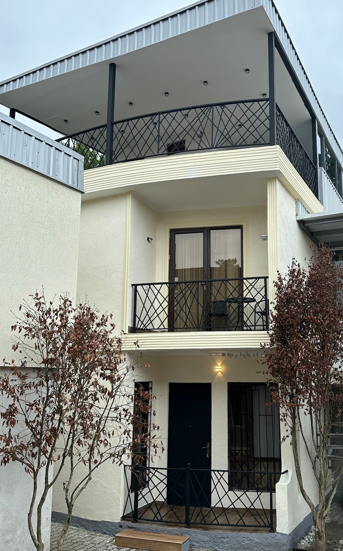 The exterior of the newly renovated apartment is captured, featuring a three-story design with a half-covered terrace on the top floor. Black railings accent the balconies, while the entrance is marked by a dark door framed by light-colored walls and bordered by small trees.