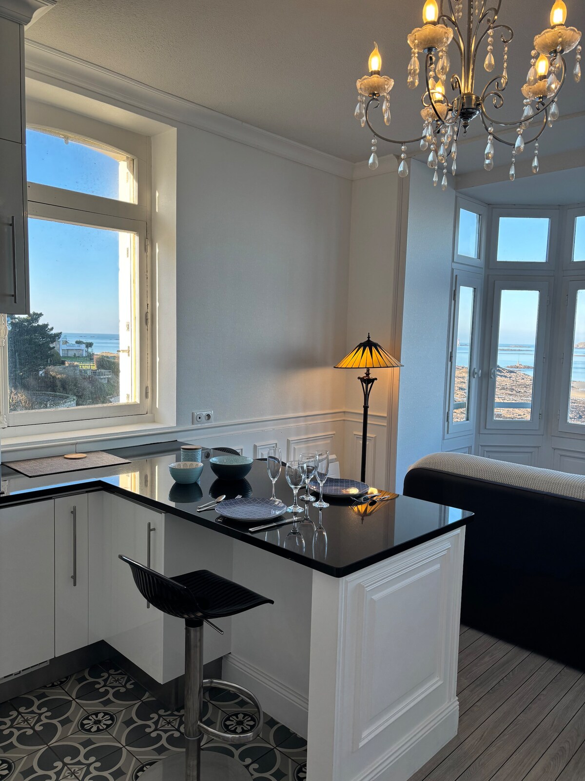 A kitchen area features a sleek black countertop with gleaming bar stools. Natural light floods in through large windows, offering a glimpse of the sea. A stylish chandelier hangs above, complementing the elegant decor.