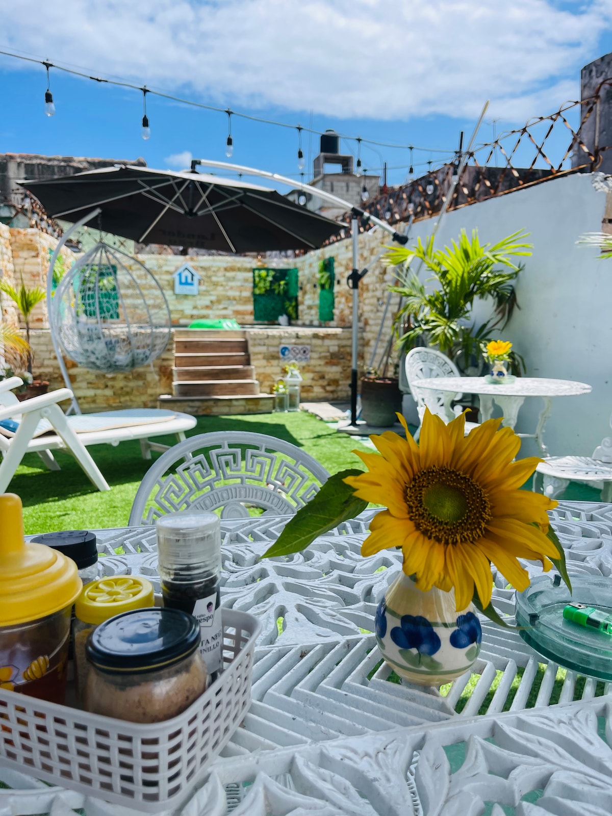 A vibrant terrace space is presented, featuring white wicker furniture and a sunny sunflower on a decorative table. The background includes steps leading to a shaded area under an umbrella, decorative greenery, and string lights overhead, contributing to a relaxing outdoor atmosphere.