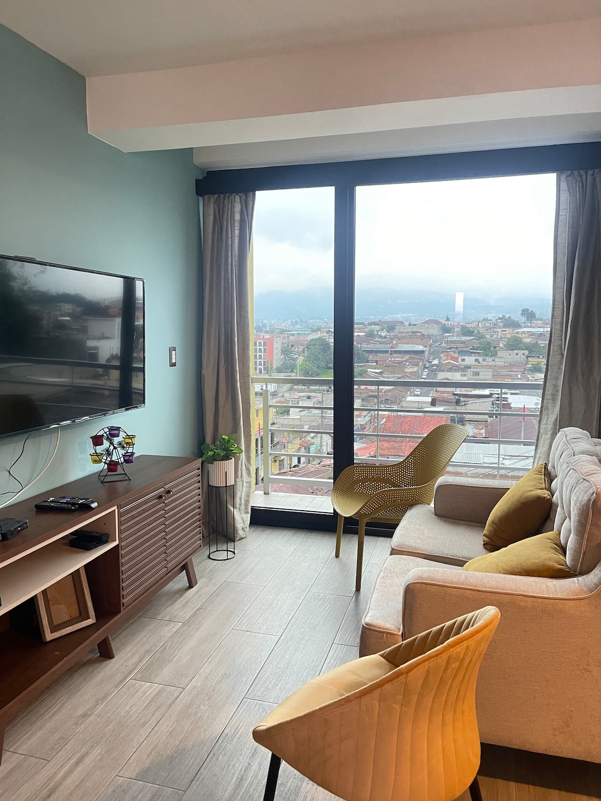 A modern living area features a beige sofa and a yellow accent chair, positioned to take in views through large glass doors. A wooden entertainment unit holds a television, while a small plant adds a touch of greenery. Natural light brightens the space.