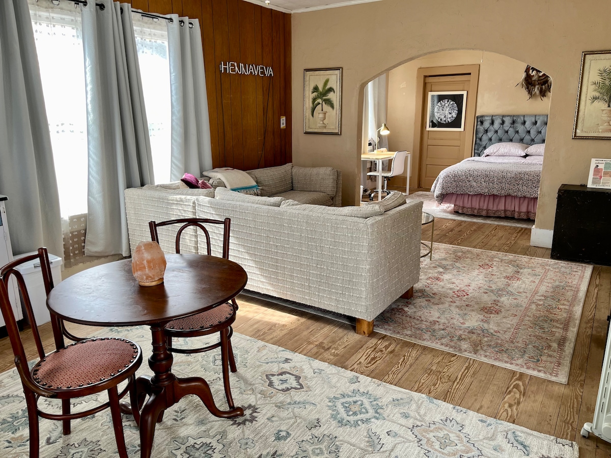 A warm interior showcases a wrap-around couch and a vintage dining table with two chairs. Soft light filters through sheer curtains, illuminating the space. A plush bed is located in the background, framed by art pieces and a calming rug underfoot.