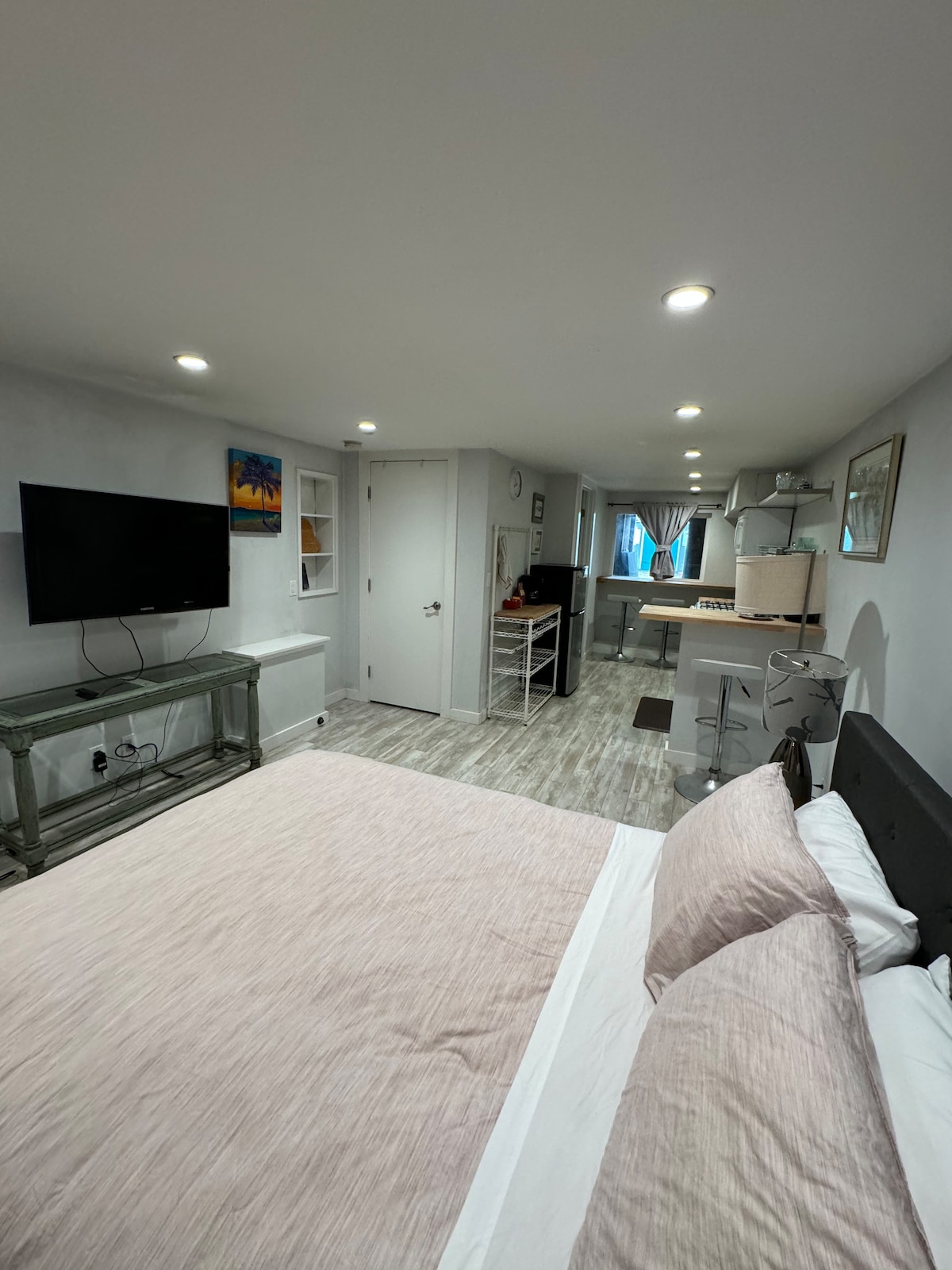 A well-lit living space features a queen bed with soft linens and a neutral color palette. A flat-screen TV is mounted on the wall, while a small kitchenette with barstool seating is visible in the background, enhancing the room's comfort and functionality.