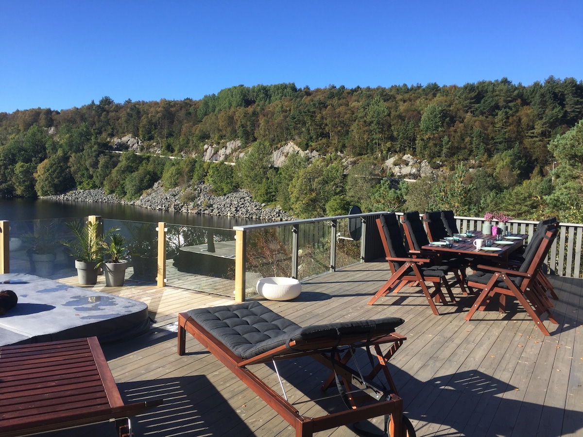 An outdoor deck offers a serene view of the surrounding nature, featuring several loungers and a dining table set for eight. Potted plants add a touch of greenery, while a hot tub is partially visible. The area is framed by trees and a calm water body.