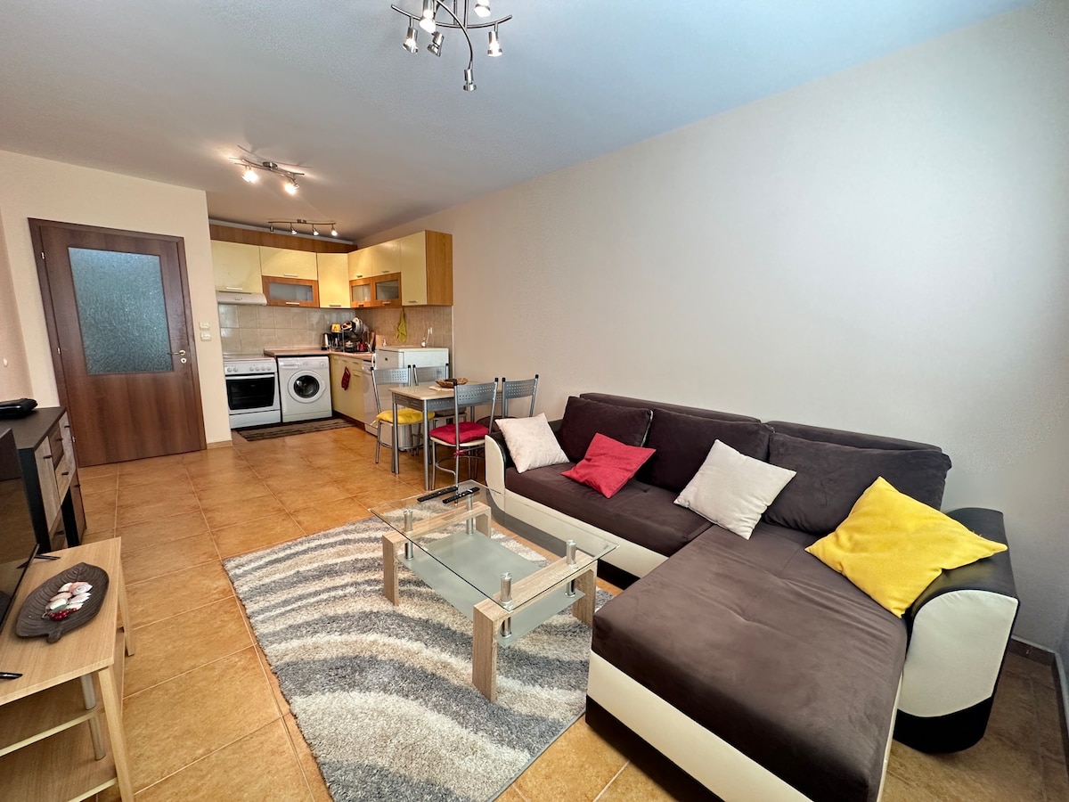 A modern living space showcases a comfortable sectional sofa with colorful cushions. A glass coffee table rests on a striped area rug, while an open kitchen with light wood cabinetry and appliances is visible in the background. Soft lighting enhances the inviting atmosphere.