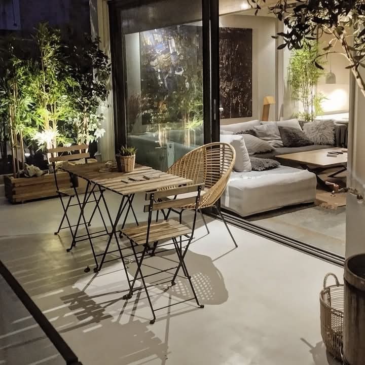 An inviting outdoor balcony area features a dining table and chairs crafted from natural materials. Lush green plants are positioned around the space, while evening lighting casts a warm glow across the furniture. Large glass doors connect this outdoor space to the interior.