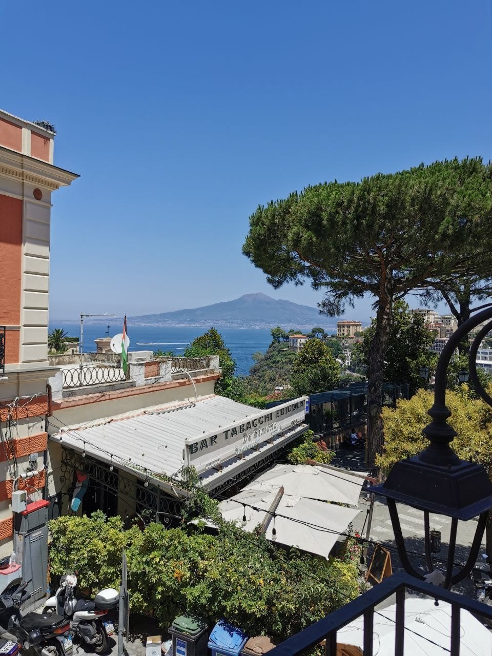 A view from the balcony reveals vibrant greenery and a bustling outdoor area below. The serene blue sea stretches out, framed by a prominent view of a distant mountain. Clear skies create a bright atmosphere, enhancing the natural beauty of the surroundings.