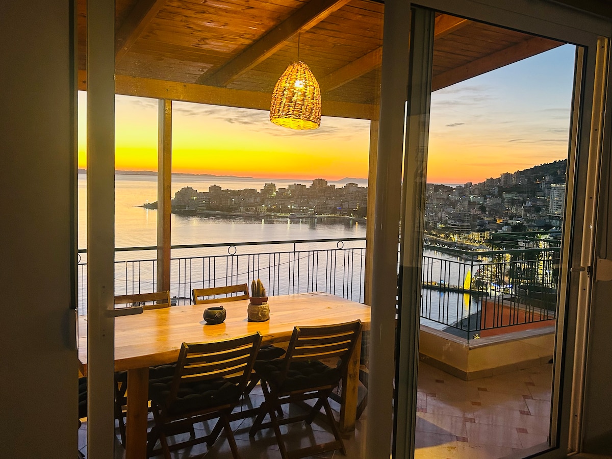 A spacious balcony dining area is depicted, showcasing a large wooden table surrounded by chairs. A warm sunset hues illuminate the scene, while views of the sea and nearby city are framed through large glass doors, highlighting the inviting atmosphere.