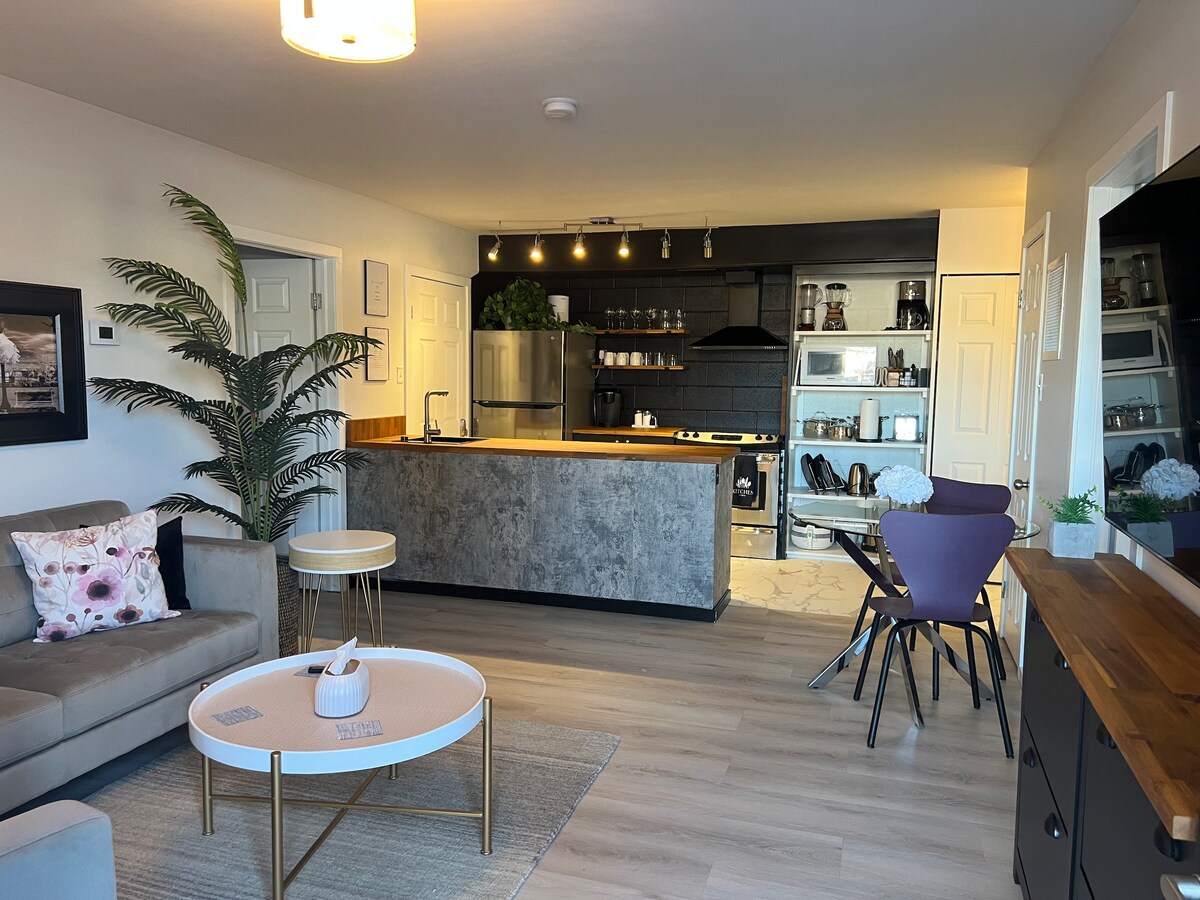 The living area features an open concept design with a cozy sofa and a round coffee table. A modern kitchen with sleek countertops and stainless-steel appliances is visible in the background, complemented by pendant lighting and a dining table with stylish chairs.
