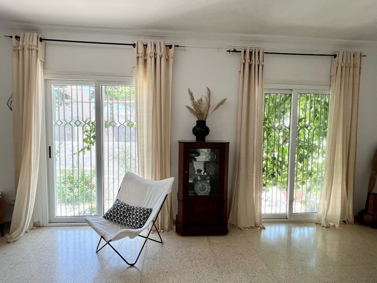 A bright living area features two large windows with white drapes, allowing natural light to fill the space. A light-colored chair is placed nearby, with a decorative plant visible in the background. A wooden cabinet displaying items stands against the wall.