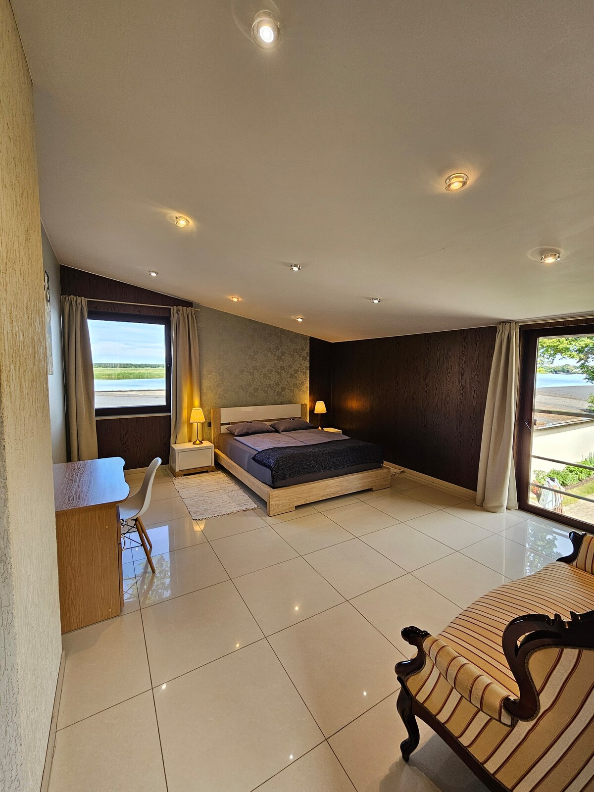 The spacious bedroom features a large bed positioned against a textured feature wall. Natural light fills the room through two windows, one of which opens onto a private terrace. A small desk and chair are placed near the entrance, complemented by a classic seating area across the room.