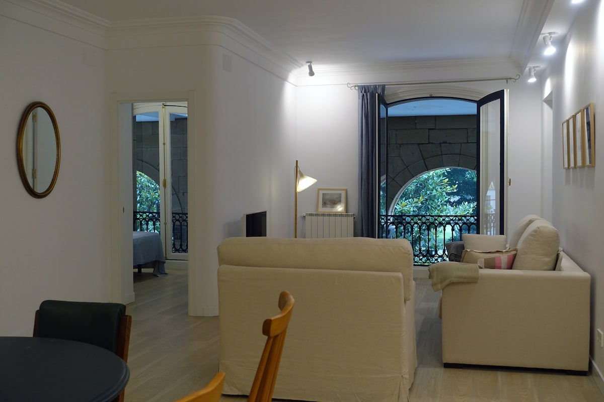 A spacious living area features large windows with views of a green tree-lined street. Light-colored furniture is positioned around a circular dining table. Soft lighting and modern decor are complemented by elegant archways leading to the bedrooms.