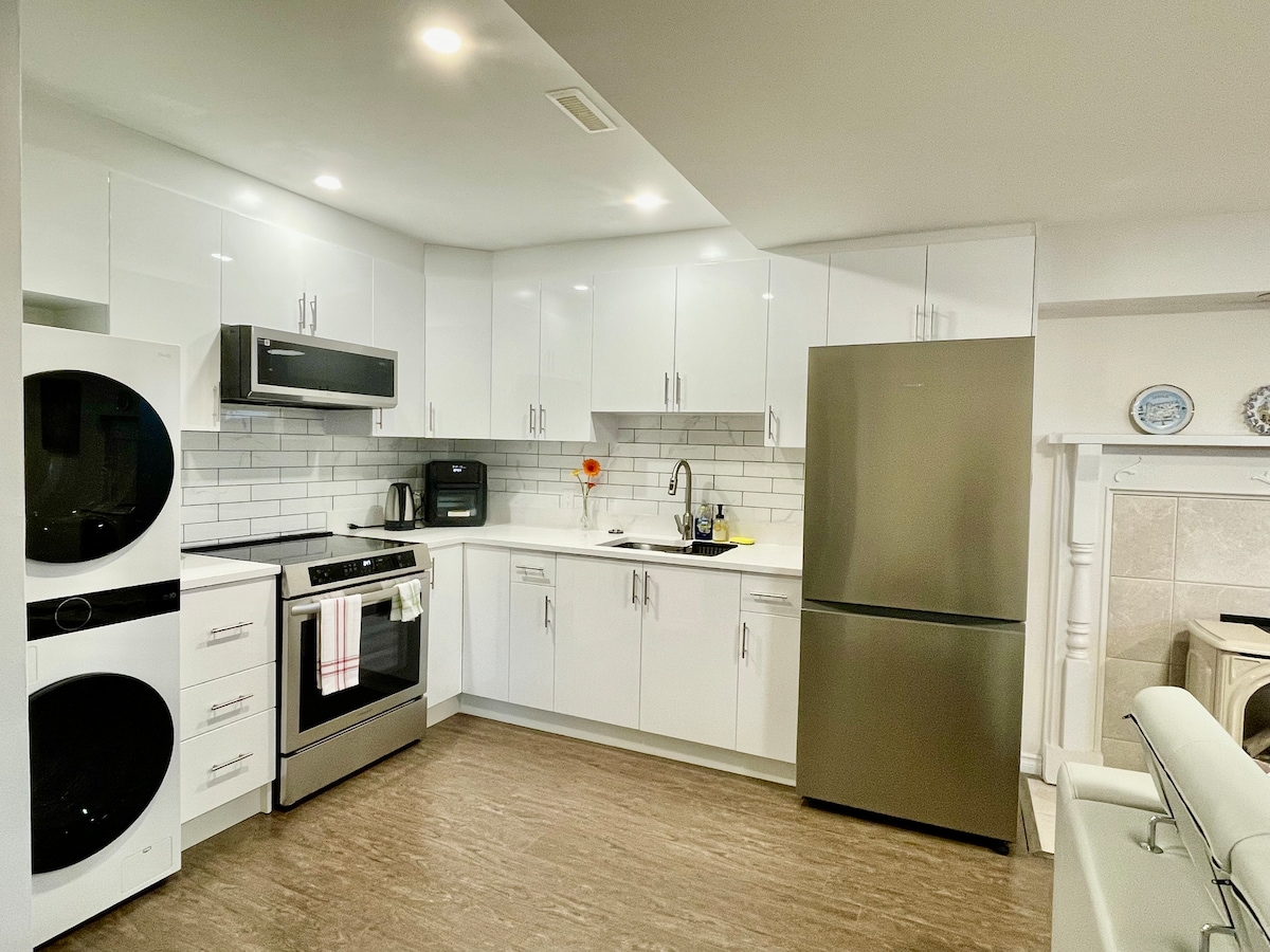 A modern kitchen features stainless steel appliances, including a refrigerator, oven, and microwave. White cabinetry and light tile backsplash create a clean and bright atmosphere. A washer and dryer are also present in the space, with subtle lighting enhancing the usability.