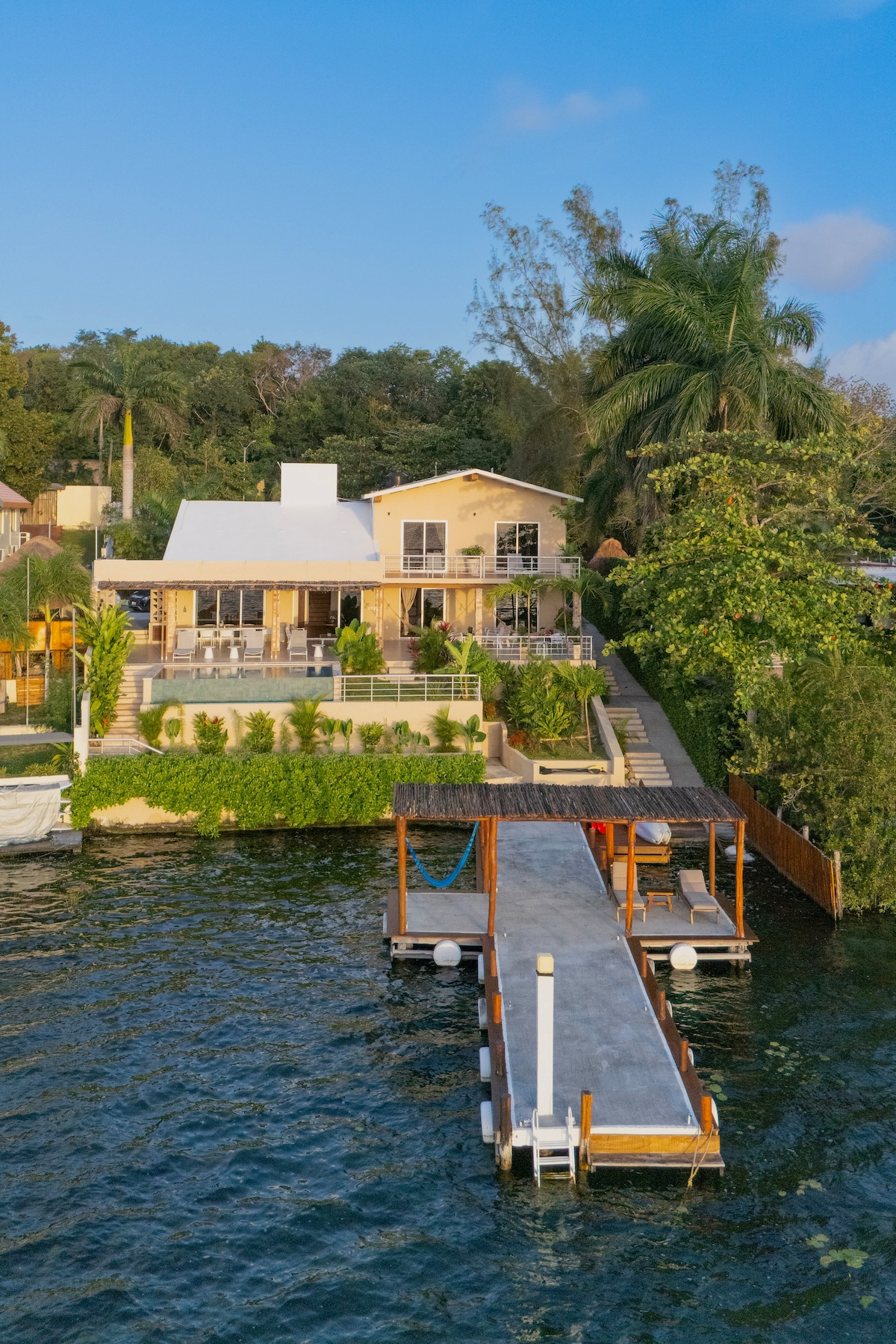 The exterior of the house is showcased against a clear blue sky, featuring a spacious terrace and lush greenery. A well-maintained dock extends into the water, supported by wooden beams, providing an inviting access point to the Cenote Esmeralda.