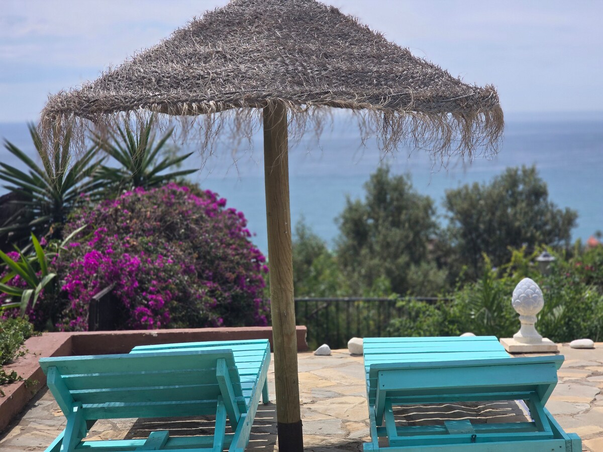 A shaded area features two turquoise loungers, positioned near vibrant pink bougainvillea and lush greenery. The tranquil sea view is visible in the background, framed by palm trees and a coastal landscape, creating a serene outdoor setting.