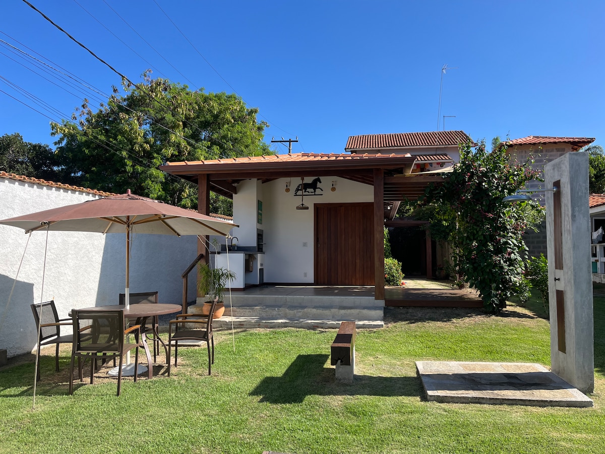 A charming guesthouse is presented with a spacious outdoor area featuring a table and chairs shaded by an umbrella. Lush grass surrounds the structure, which showcases a wooden entrance and large windows, inviting natural light into the airy interior.