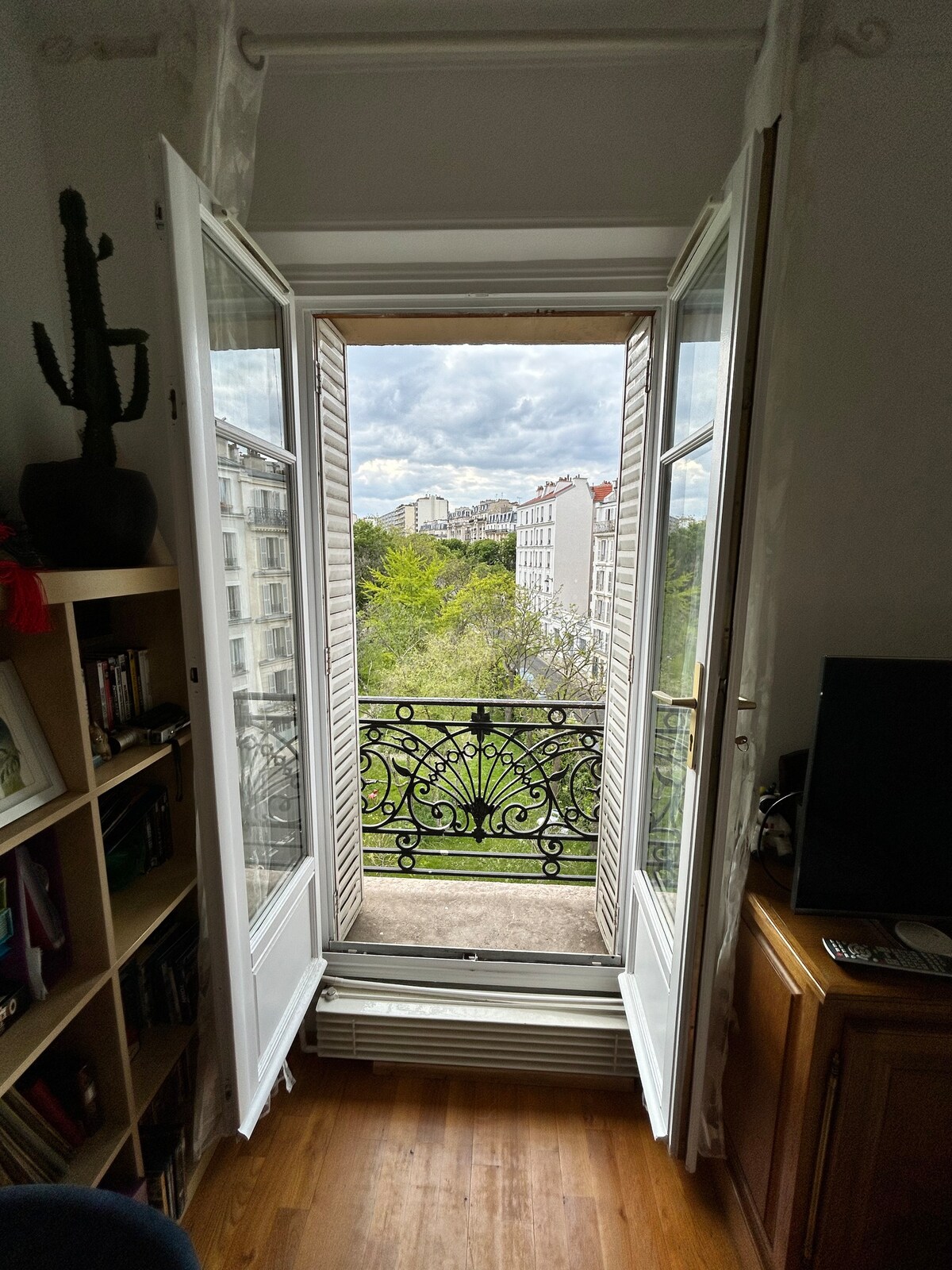 The image shows double doors opening to a view of a green courtyard, framed by ornate black railings. Natural light fills the space, highlighting the warm wooden flooring. Nearby, a bookshelf is visible, alongside a wood cabinet and a television.