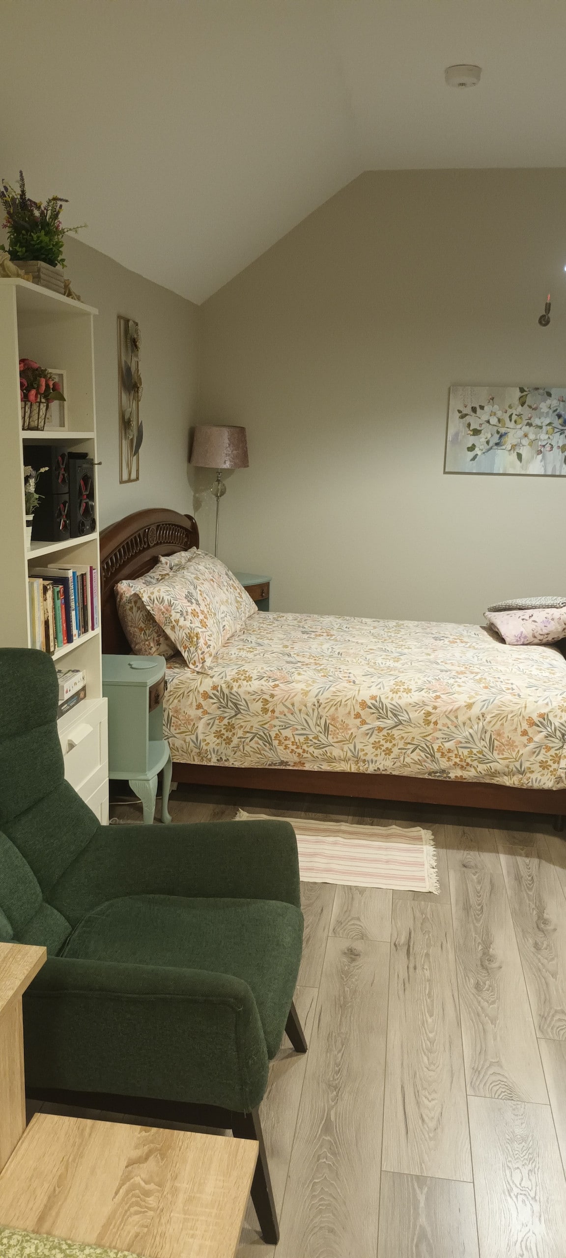 An open plan sleeping area features a cozy bed adorned with a floral-patterned coverlet. A small nightstand with a lamp is positioned beside the bed, and a comfortable green chair is placed nearby. Shelves filled with books enhance the welcoming atmosphere.
