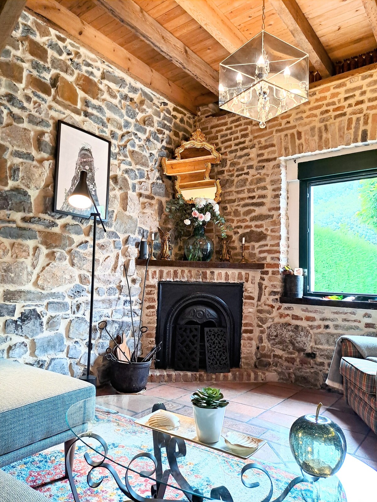 A living room corner features a stone wall, a charming fireplace, and a decorative wooden shelf adorned with flowers. Natural light enters through a large window, while a cozy seating area is complemented by a colorful rug and minimal decorative items.