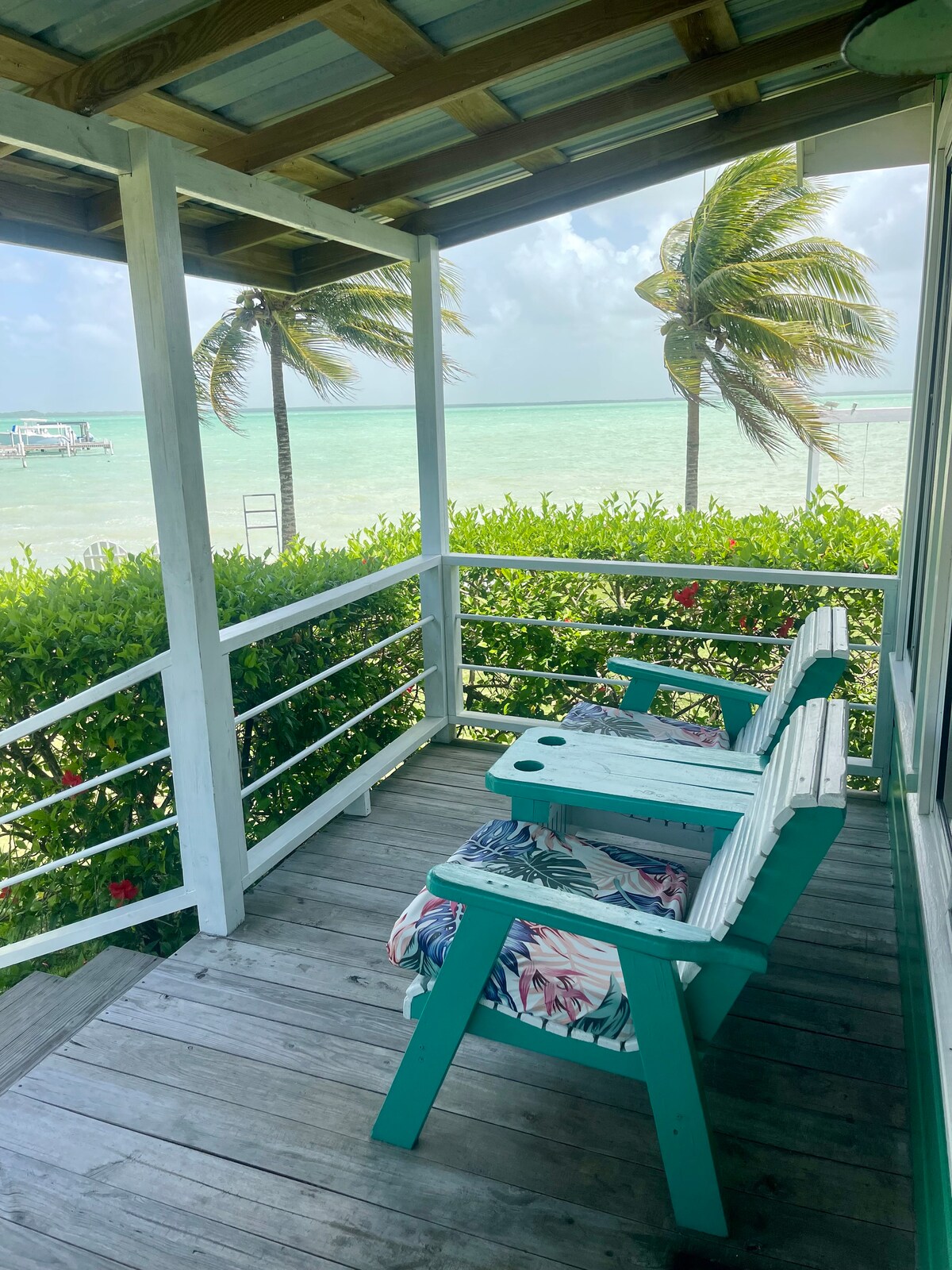 A covered porch area features two teal Adirondack chairs, suggesting a relaxed outdoor seating option. Lush greenery and tropical plants surround the space, while palm trees sway gently in the breeze. The turquoise waters of Corozal Bay are visible in the background.