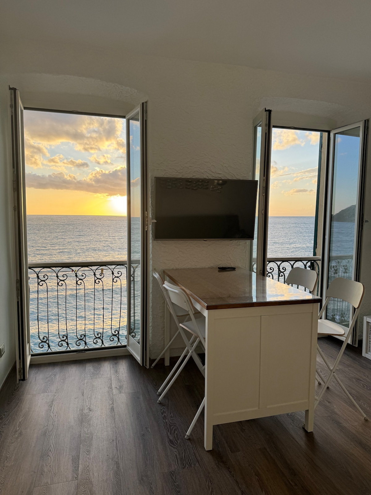 An inviting dining area is situated near two open doors, revealing a scenic sea view. A small, light wooden table is accompanied by two white chairs. A flat-screen television is mounted on the wall, complementing the bright atmosphere created by the sunset reflected on the water.