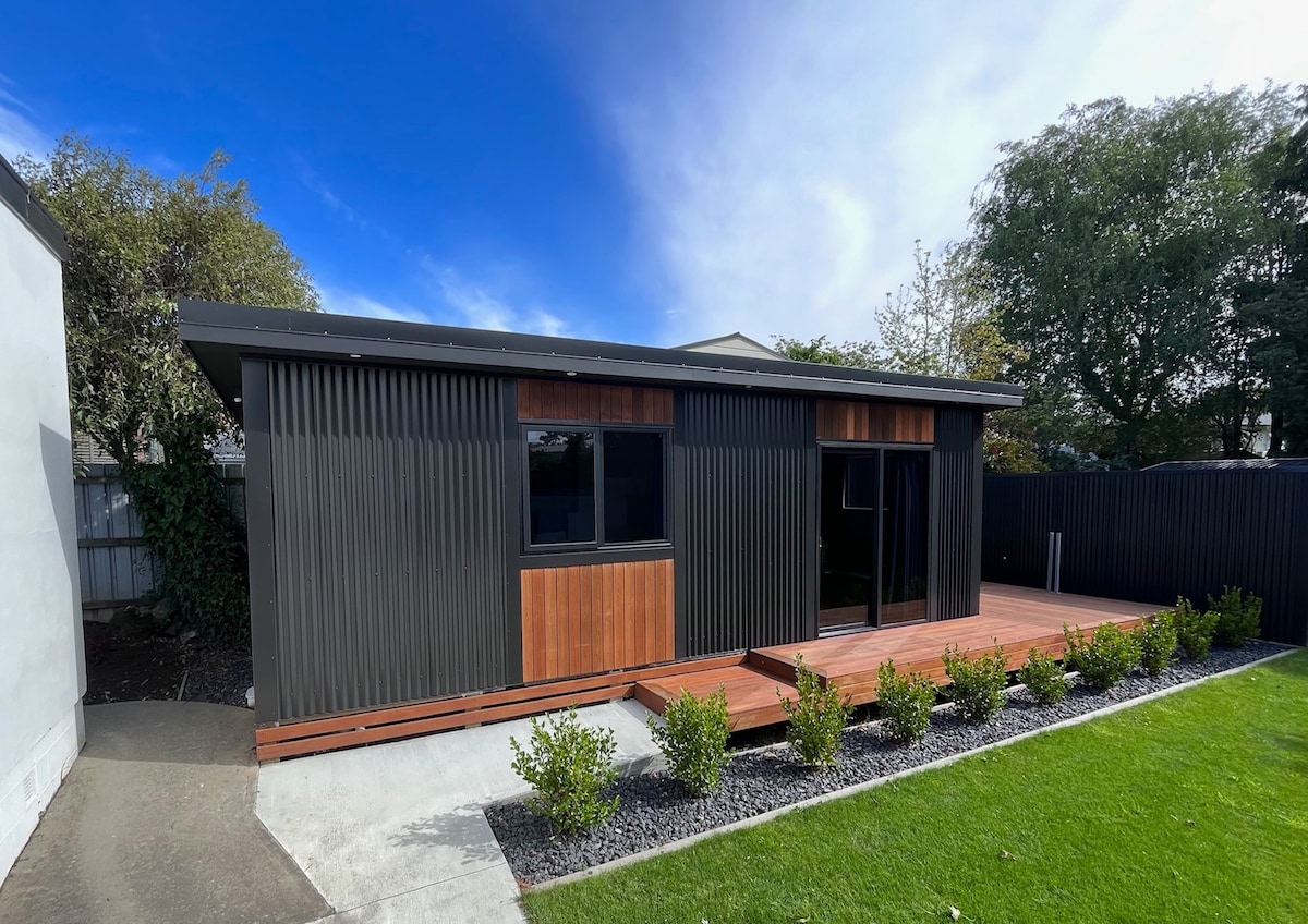 A modern tiny home is presented with a sleek metallic exterior, accented by wooden panels. The structure features large windows, allowing natural light to fill the space. A wooden deck leads to the entrance, surrounded by neatly trimmed shrubs and a grassy area.